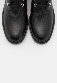 Czarne skórzane buty o teksturowanym wykończeniu, zaokrąglone noski i metalowe oczka, z czarnymi sznurowadłami oraz kontrastującym przeszyciem wzdłuż podeszwy.