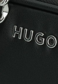 Materiale nero testurizzato con lettering "HUGO" argentato. Dettaglio hardware prominente. Inquadratura ravvicinata che mette in evidenza design e finitura.