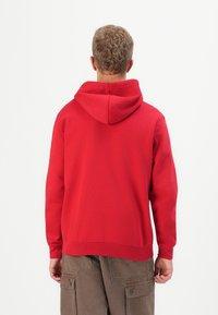 Man met kort, krullend haar in een rode hoodie en een bruine cargo broek, gezien van achteren tegen een plain witte achtergrond.