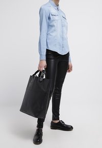 Black leather tote bag with double handles, sleek shape, and front pocket; paired with light blue button-up shirt and black leather pants.