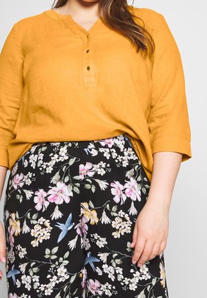 Femme portant un haut moutarde boutonné à manches trois-quarts et un pantalon noir avec des motifs floraux et d'oiseaux colorés.