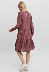 Abito lungo a maniche, in tessuto malva a balze. Presenta un dettaglio a chiave nella parte posteriore e una silhouette rilassata, accentuata da un materiale morbido e fluente.