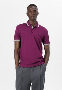 Lila Poloshirt mit kontrastierendem gestreiften Kragen und Bündchen, mit kleinem schwarzem Logo. Modell trägt graue Hose, Hände in den Taschen.