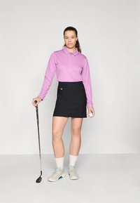 Fioletowa koszulka polo z długim rękawem na zamek, zestawiona z czarną spódnicą. Szare buty z jasnymi skarpetkami, trzymająca kij golfowy i piłkę.