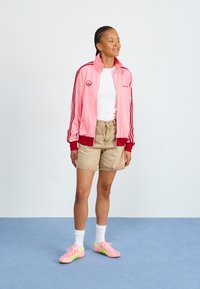 Rózsaszín zipzáros sportdzseki piros díszítéssel, fehér póló, bézs rövidnadrág és rózsaszín cipő zöld csíkkal egy kék padlón.