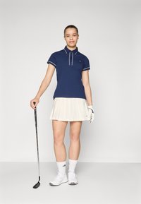 Marineblauw polo met korte mouwen en witte accenten, gecombineerd met een geplooide crèmekleurige rok. Houdt een golfclub vast, draagt witte schoenen en sokken.
