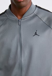 Detailný záber na osobu oblečenú v šedej zipsovej bunde s čiernym logom Jumpman na ľavej hrudi a čiernymi pruhmi na pleciach.
