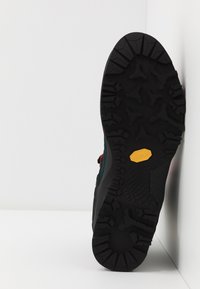 Svart Vibram-såle med dype mønstre, flerfarget overdel med teksturert stoff og en gul logoaccent. Sidevisning mot en hvit bakgrunn.