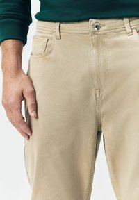 Man wearing beige pants and green long-sleeve shirt, hand resting on thigh, visible button and pocket details on pants.