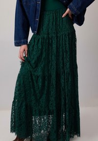 Gonna maxi verde in pizzo con un design a strati e motivi floreali testurizzati. Abbinata a una giacca di jeans, con una mano appoggiata sulla gonna.