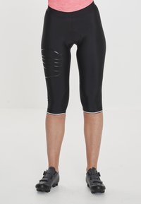 Schwarze Radsportshorts mit enger Passform, ausgestattet mit einem strukturierten seitlichen Akzent und knielangem Schnitt, kombiniert mit schwarzen Radschuhen.