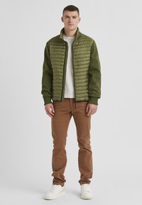 Grüne Pufferjacke mit gerippten Ärmeln, hohem Kragen und Reißverschluss, kombiniert mit braunen Hosen und weißen Sneakers.