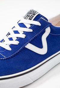 Sininen mokkanahkakengät valkoisilla nauhoilla, valkoinen aalto-logo sivussa, ja "Vans Off The Wall" -labeli kielen päällä, pohjan yläpuolella mustalla raidalla.