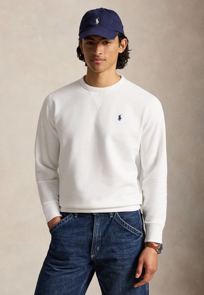 Ung mand iført en hvid sweatshirt med en blåt broderet logo, mørkeblå jeans, en navy kasket og et armbåndsur, stående med hænderne i lommerne.