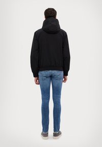 Black hooded jacket with a relaxed fit, elastic waistband, and long sleeves, paired with blue jeans and gray sneakers, shown from the back.