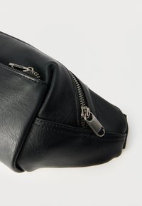 Μαύρη δερμάτινη τσάντα με ομαλή υφή, που διαθέτει τσέπη με φερμουάρ και ασημένια μεταλλικά στοιχεία. Ο σχεδιασμός είναι συμπαγής με διακριτική καμπυλότητα.