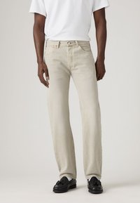 Man wearing beige straight-leg jeans, white t-shirt, white socks, and black tassel loafers, standing against a plain light background.