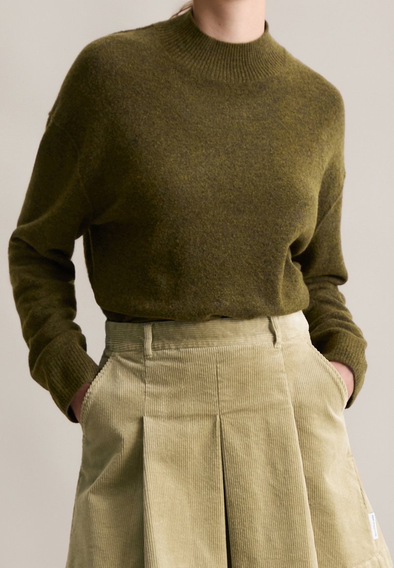 Personne portant un pull en maille vert olive rentré dans une jupe en velours côtelé beige taille haute, les mains dans les poches.