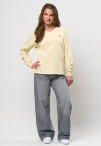 Jonge vrouw staand met hand op heup, gekleed in een crèmekleurige longsleeve, losse grijze spijkerbroek en witte sneakers tegen een witte achtergrond.