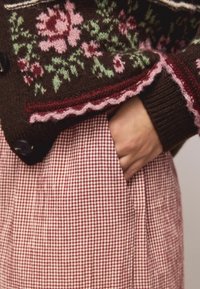 Cardigan maro tricotat cu modele florale în roz și verde, asortat cu o fustă în carouri roșu și alb. Detaliu cu mâna în buzunar vizibil.