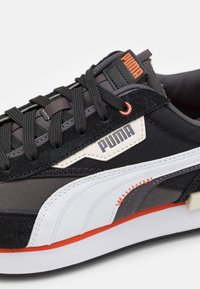 Sneaker in tessuto nero con accenti grigi e bianchi, caratterizzato da un motivo testurizzato, cuciture a contrasto e un logo Puma prominente sul lato.