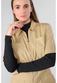 Une combinaison zippée couleur tan avec des manches courtes, dotée de deux poches sur la poitrine. Associée à un pull noir à côtes avec des accents métalliques.