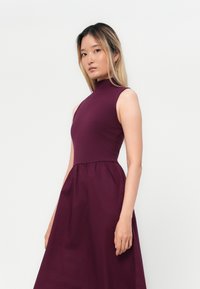 Burgundin mekko, jossa on korkea pääntie, hihaton muotoilu ja rypistetty hame. Valmistettu sileästä kankaasta, jossa on yhtenäinen väri.