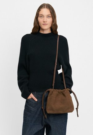 Donna che indossa un maglione di lana nero e jeans scuri, mentre tiene una borsa a spalla in suede marrone con manici in pelle.