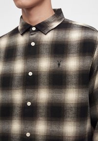 Homme portant une chemise à carreaux noire et beige boutonnée, avec un petit logo brodé sur la poitrine, col visible, torse supérieur montré.