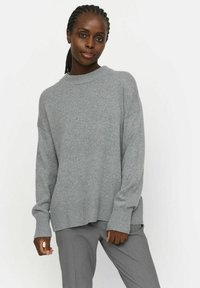 Grauer Strickpullover mit lockerem Schnitt, gerippten Bündchen und Saum. Weiche Textur, weitem Ausschnitt und leicht längerer Rückseite. Kombiniert mit grauen Hosen.