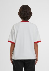 T-shirt bianco morbido con maniche corte, colletto rosso e polsini rossi. Texture liscia e design casual; etichetta visibile di lato.