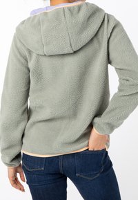 Chaqueta de forro polar suave y verde claro con capucha, que presenta una textura lisa, un dobladillo bordeado en melocotón y puños ajustados.