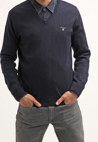 Granatowy sweter typu v-neck z dzianiny, z małym logo na piersi. Nosi go w zestawie z dżinsową koszulą pod spodem.