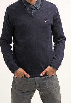 Marineblauer V-Ausschnitt-Pullover aus Strickstoff, mit einem kleinen Logo auf der Brust. Kombiniert mit einem darunter liegenden Jeanshemd.