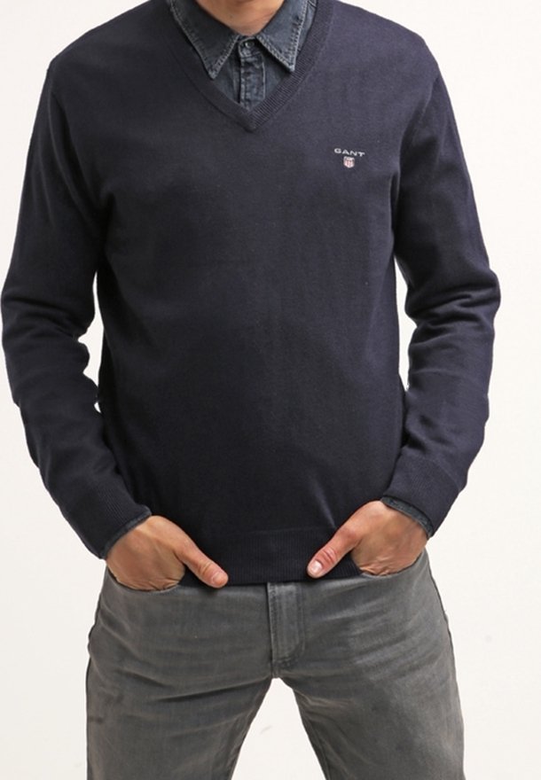 Granatowy sweter typu v-neck z dzianiny, z małym logo na piersi. Nosi go w zestawie z dżinsową koszulą pod spodem.
