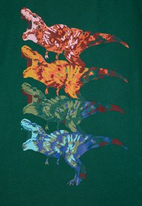 Grafisk t-skjorte med fire fargerike, pikselerte T-Rex-design i oransje, gult, grønt og blått. Bakgrunnen er mørkegrønn stoff.