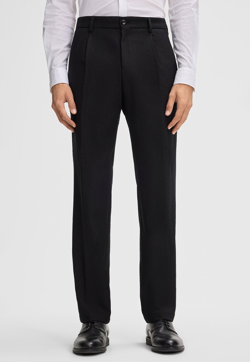 Schwarze Hosen aus strukturiertem Stoff, mit einem Knopfverschluss, zwei vorderen Taschen und einem geraden Bein Design. Getragen mit einem weißen Hemd.