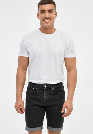 Joven sonriente, lleva una camiseta blanca lisa y pantalones cortos de mezclilla negros remangados, de pie sobre un fondo blanco.