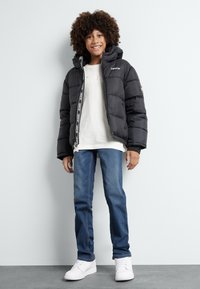 Doudoune noire avec capuche, fermeture éclair et détail de logo. Portée sur un t-shirt blanc et un jean bleu. Des baskets blanches complètent la tenue.