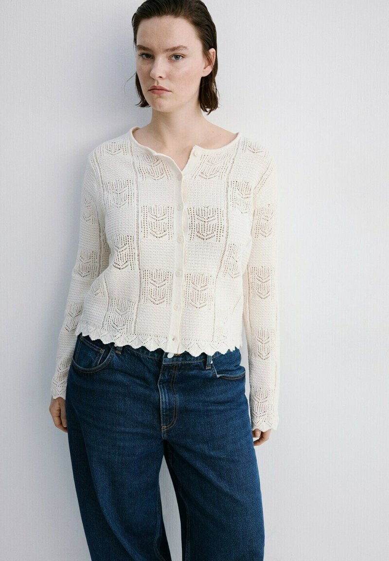Femme aux cheveux courts portant un cardigan en maille blanche à motifs et un jean bleu foncé, se tenant contre un mur blanc.