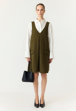 Femme portant une chemise blanche à manches longues sous une robe sans manches vert olive, tenant un sac à main noir et portant des talons noirs, debout sur fond blanc.