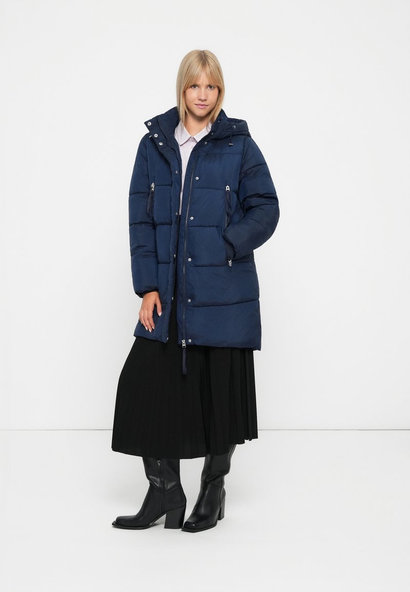 Giacca a vento blu navy con cappuccio, chiusura con zip e due tasche. Caratterizzata da sezioni trapuntate e cordoncino sull'orlo. Abbinata a una gonna nera.