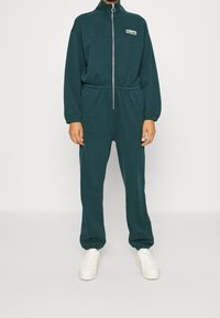 Dunkelgrüner, zipbarer Overall mit hohem Kragen, elastischem Bund und elastischen Bündchen an Ärmeln und Knöcheln. Weißes Logo auf der Brust, kombiniert mit weißen Sneakern.