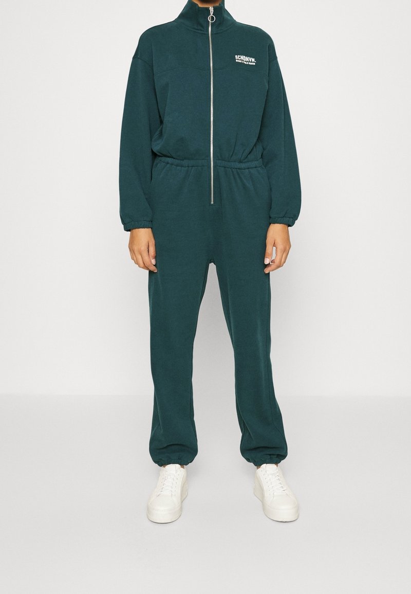 Dunkelgrüner, zipbarer Overall mit hohem Kragen, elastischem Bund und elastischen Bündchen an Ärmeln und Knöcheln. Weißes Logo auf der Brust, kombiniert mit weißen Sneakern.