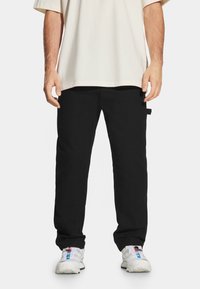 Czarne spodnie o prostym kroju, z dwiema kieszeniami bocznymi i metką na lewym biodrze. Ubrane z luźną beżową koszulką i białymi trampkami.