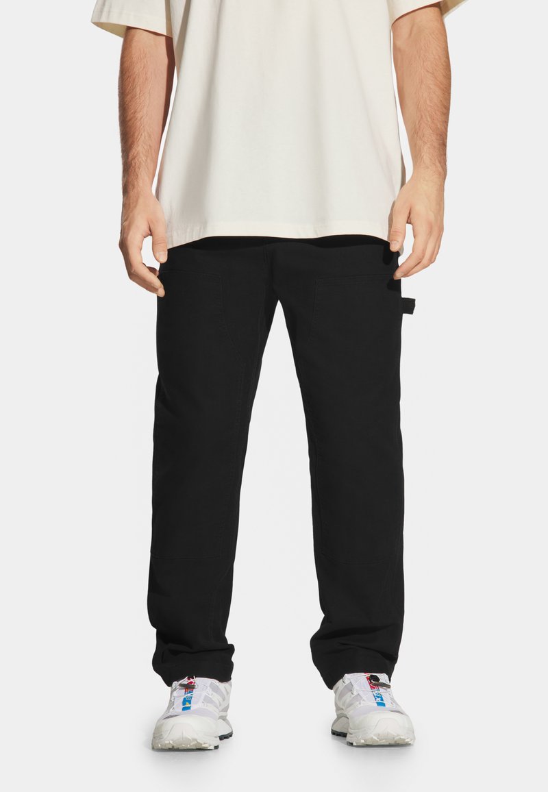 Czarne spodnie o prostym kroju, z dwiema kieszeniami bocznymi i metką na lewym biodrze. Ubrane z luźną beżową koszulką i białymi trampkami.