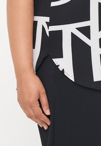 Czarny top o fakturowanej powierzchni z wyrazistym białym geometrycznym wzorem, zestawiony z gładkim czarnym dołem. Ręka delikatnie dotyka materiału.