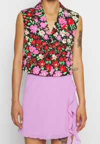 Bloemige cropped vest met een zwarte achtergrond voorzien van roze, rode en groene bloemen; gecombineerd met een lila roezelrok. Meerdere knopen zichtbaar.