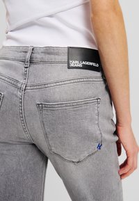 Pilkos džinsinės kelnės su juodu odiniu etiketė, ant kurios užrašyta "KARL LAGERFELD JEANS" ir mėlynas akcento logotipas. Išskirtinė siuvimo ir kišenių detalė.
