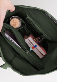 Interno di una borsa a tracolla verde con un compartimento con cerniera, una bottiglia d'acqua in bambù, un laptop e due libri, con un tirante della cerniera colorato visibile.